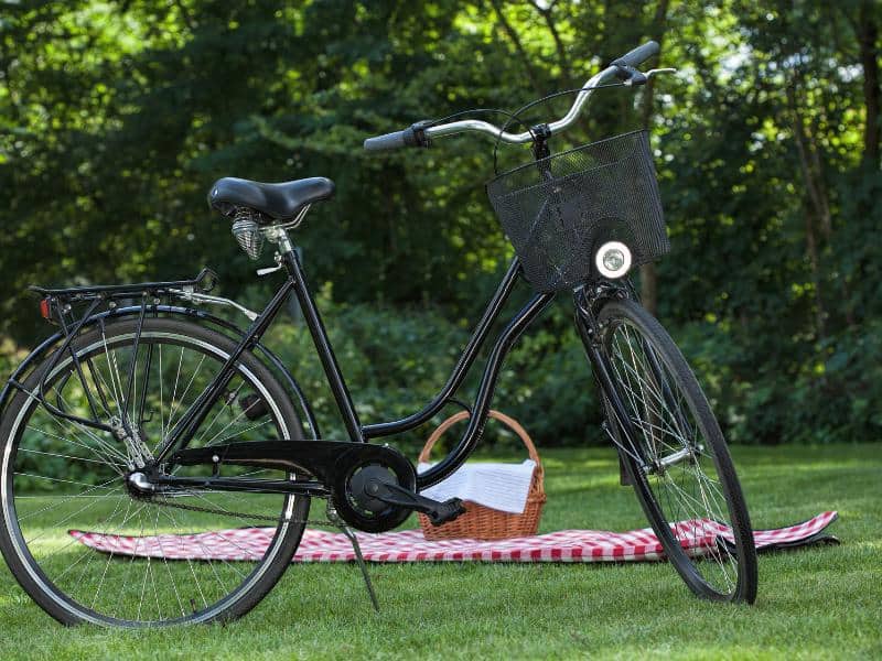 A bike next to a picnic basket on a tablecloth on the grass.