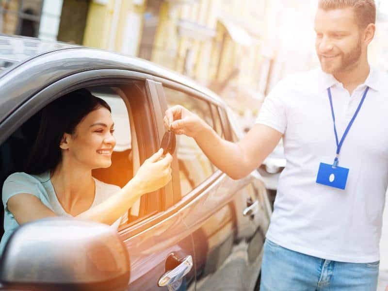 Man handing over the keys to a woman in the driver's seat who has just rented a car.