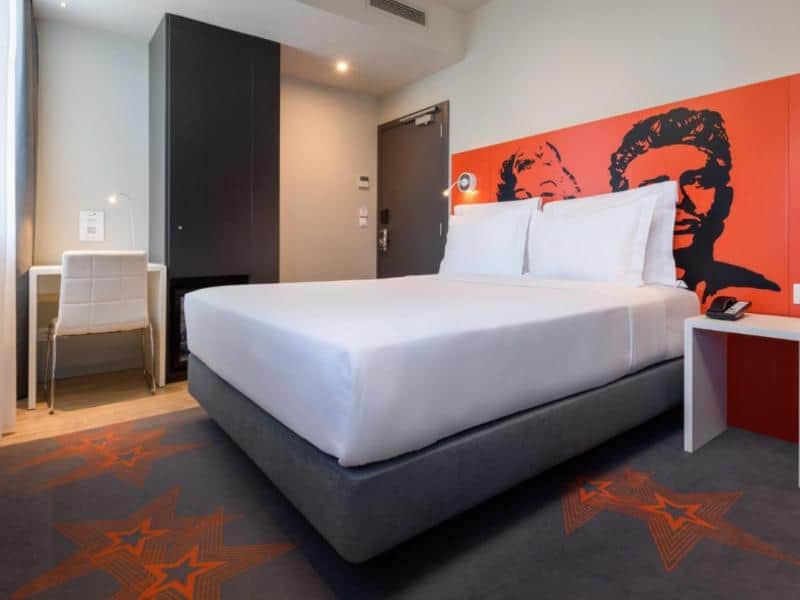 A spacious hotel room with a double bed, plush white linens, and decorative pillows. The room features a warm-toned accent wall, a seating area with an orange armchair, and a desk with a coffee machine. Natural light pours in from a large window with a city view. This inviting room is a fantastic option for anyone seeking a relaxing Lisbon airport hotel.