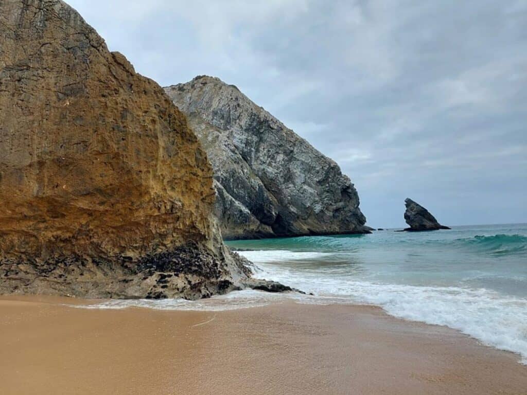 Praia da Adraga on a cloudy day has beautiful fine sand an huge boulders jutting out from the sand and water.