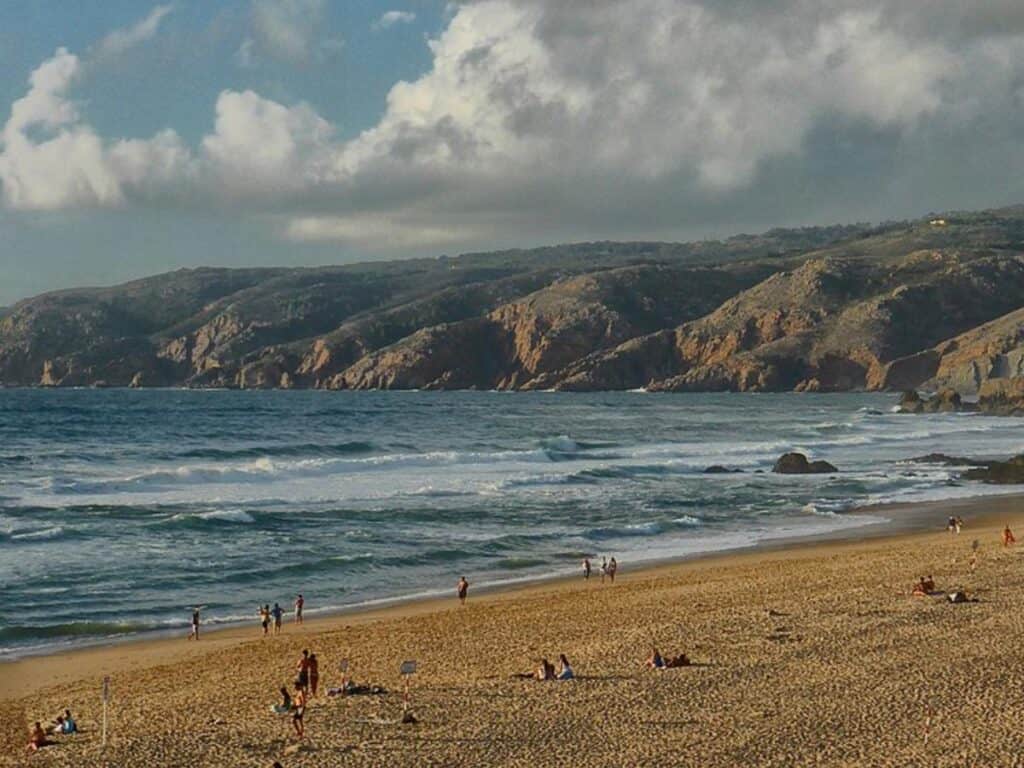 Praia do Guincho on a cloudy day with haze up on the cliffs in the background and few people on the beach.