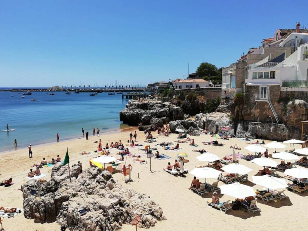 How far is Lisbon from the beach? Praia da Rainha in Cascais is a short 30 minute drive from Lisbon. Beautiful small beach  covered in patrons under sunshades with homes built into the cliff to the right and the dock on the horizon.