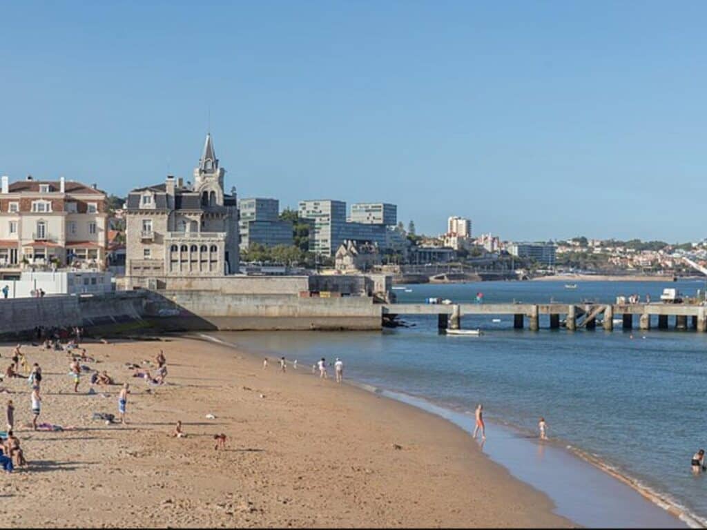 Several people mulling around in bathing suits on Praia da Ribeira de Cascais and Cascais in the background on a sunny day.