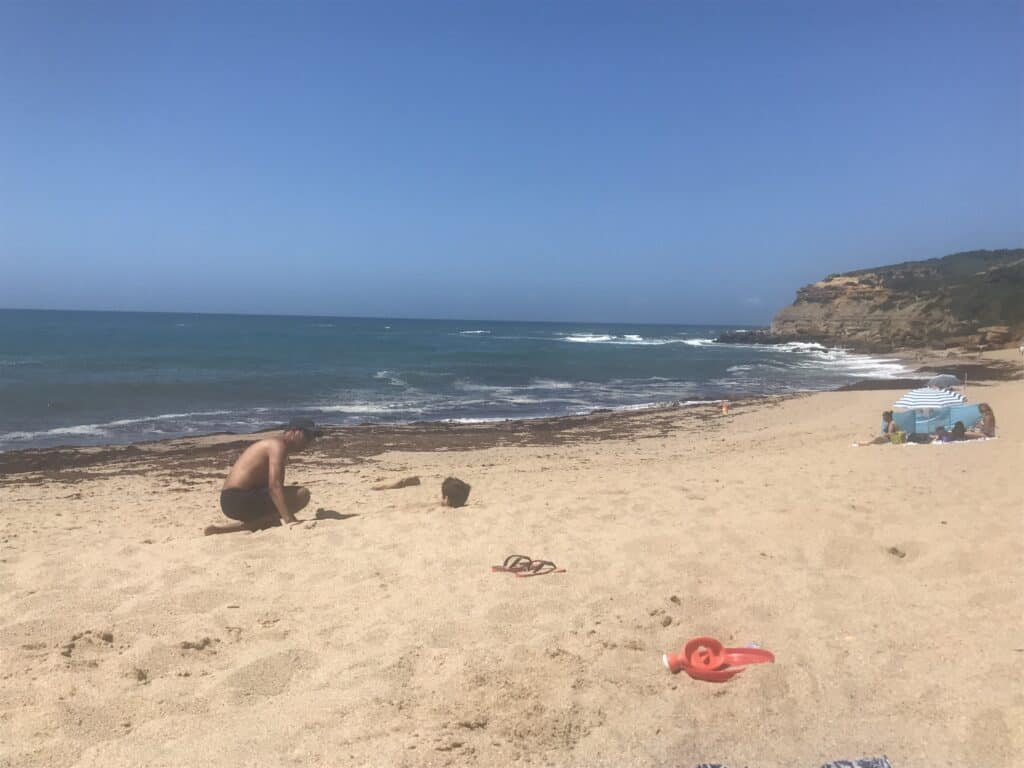 Beach in Portugal on a sunny day, with cliffs in the background to the right, seaweed on hte shore, and a striped blue umbrella on the beach next to a man burying a boy in the sand.