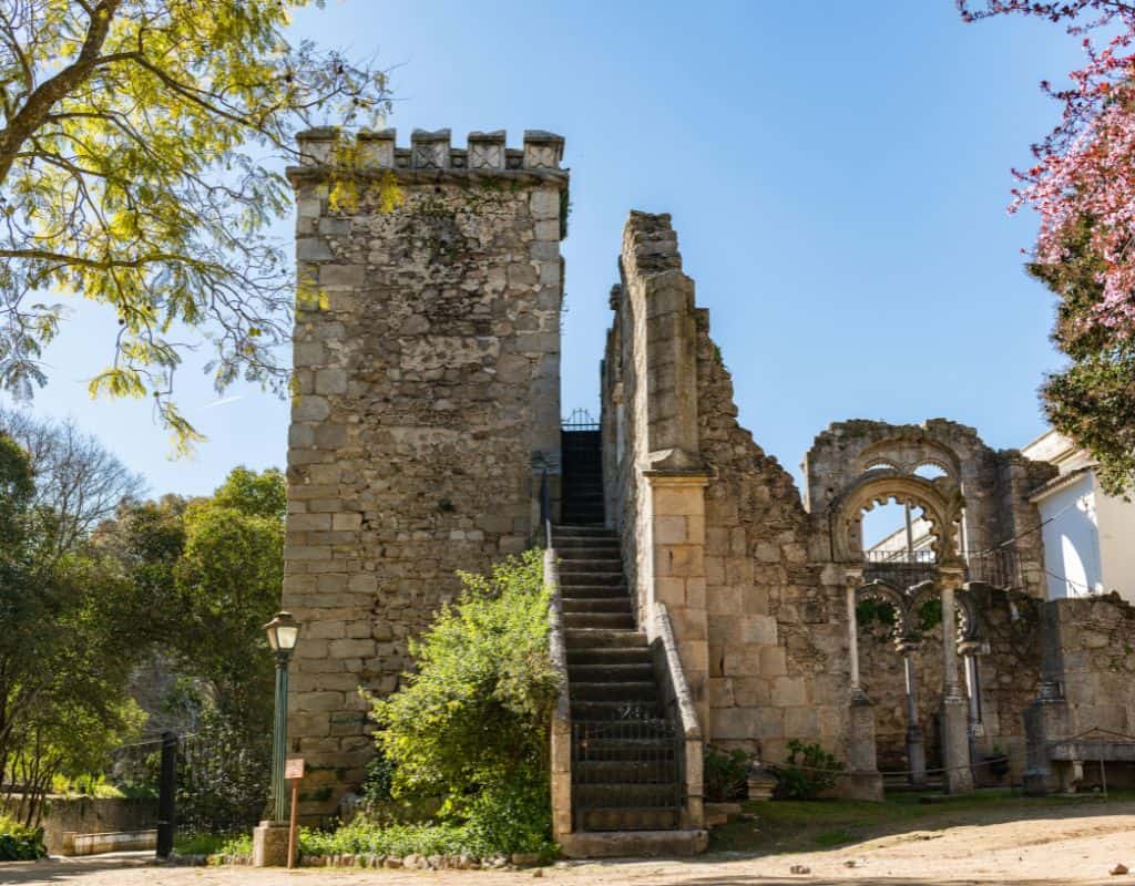 The collection of stairwell, windows and pillars found around Evora which have been joined in the Evora Public Gardens as a conversation piece. Beautiful pink flowers on the right, and green trees on the left.