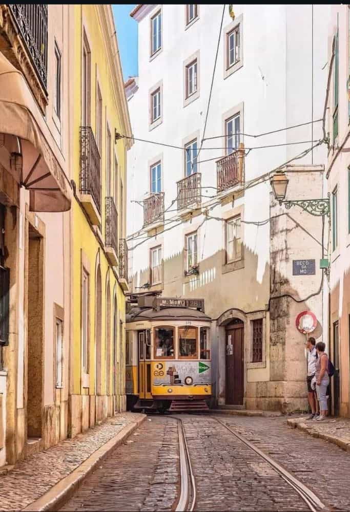 A yellow tram navigating a narrow street in Lisbon, Portugal, lined with pastel-colored buildings. The street is paved with cobblestones, and two people stand near a building on the right side. The tram is marked with the number "28E" and the destination "M. Moniz."