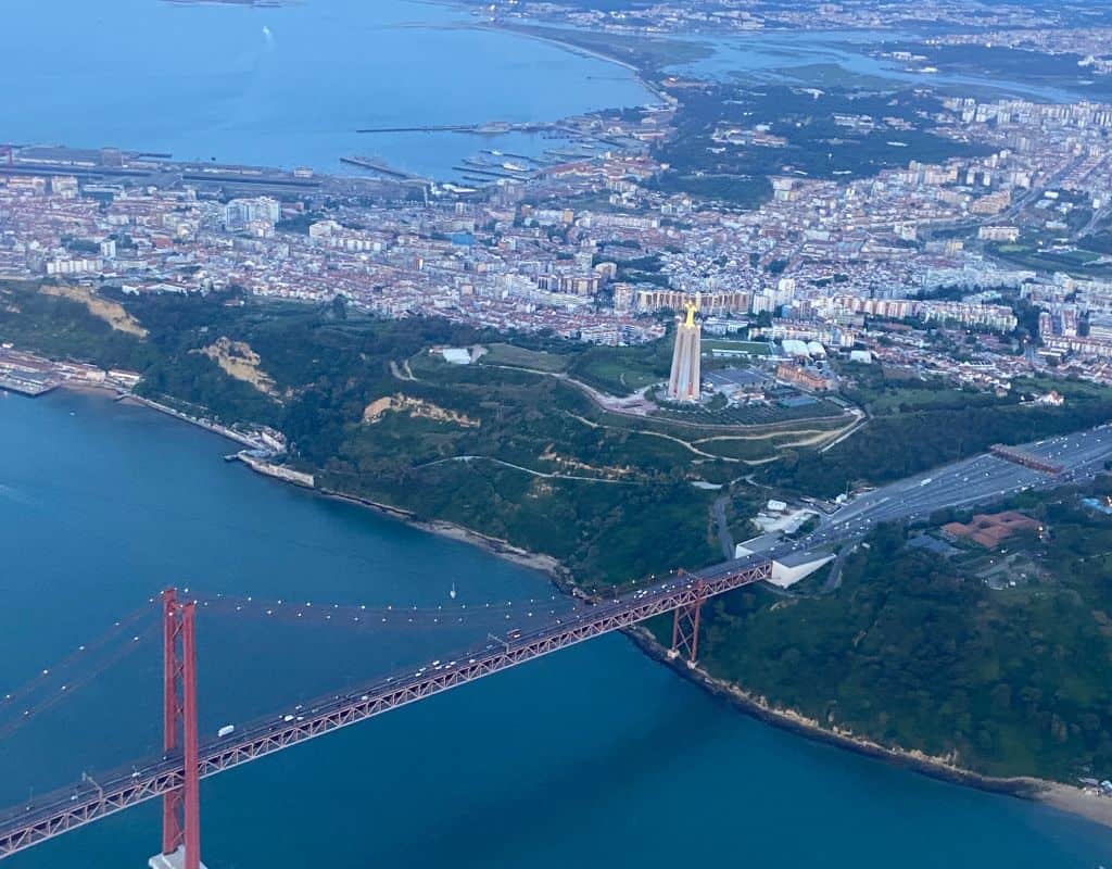 An aerial view of Lisbon, Portugal, featuring the iconic 25 de Abril Bridge and the Christ the King statue. The city sprawls out in the background, bordered by the blue waters of the Tagus River.