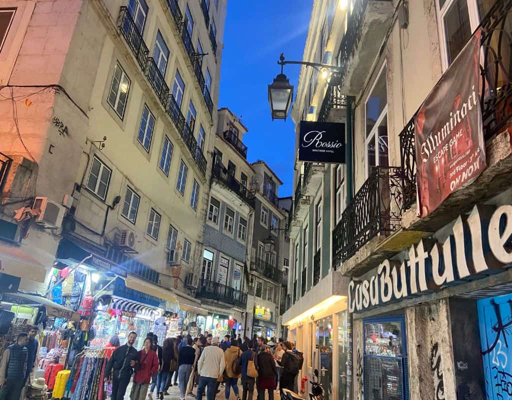 A vibrant street in downtown Lisbon, Portugal, at night. The street is crowded with people and lined with shops, cafes, and signs, creating a lively, energetic atmosphere under the evening sky.
