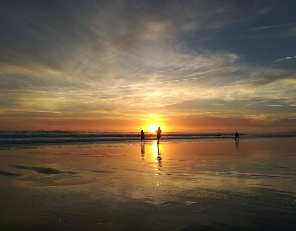 A serene beach at sunset on Carcavelos beach in Lisbon, Portugal. The sky is filled with warm hues of orange, yellow, and purple, reflecting on the wet sand. Silhouettes of people are visible walking and playing in the distance. Make sure to go to the beach if you spend 4 days in Lisbon in summer.