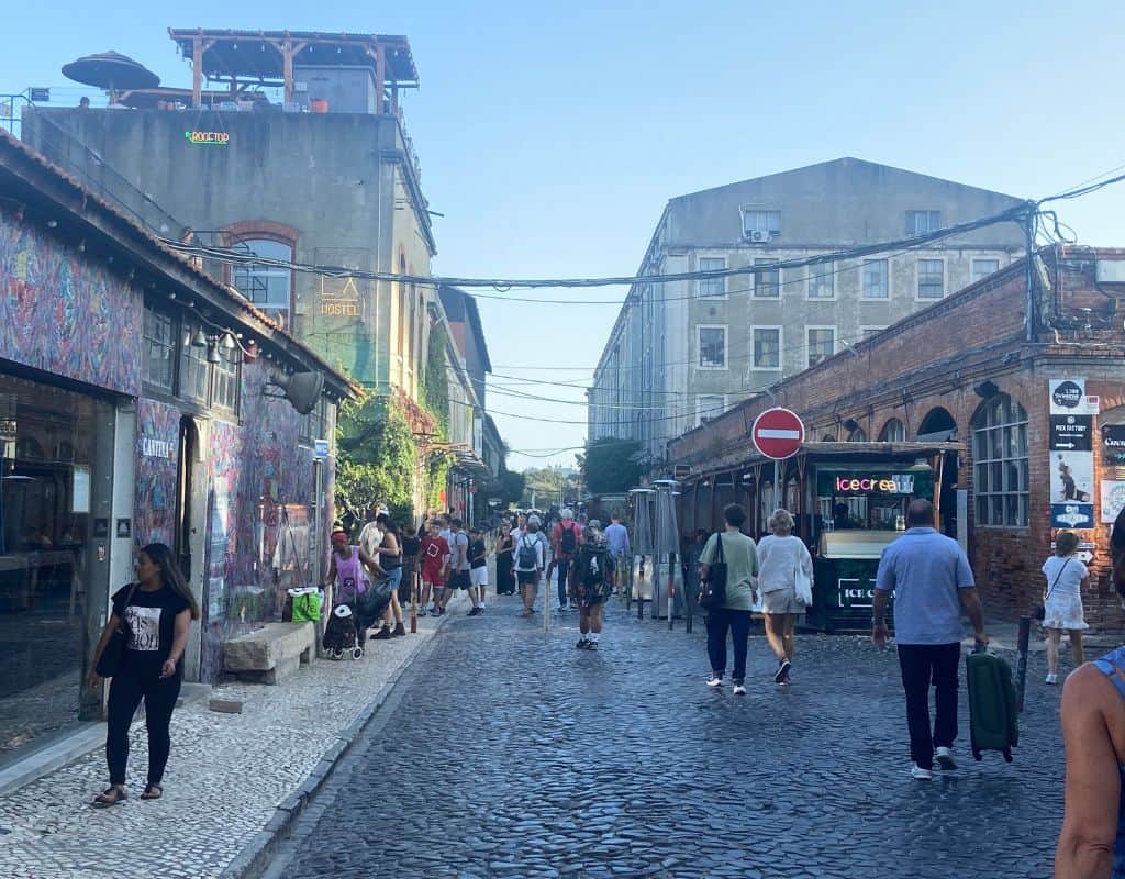 A bustling street in Lisbon, Portugal, with people walking on a cobblestone road. Buildings with colorful graffiti and signs for a hostel and ice cream shop line the sides. The street is located in LXFactory and has a lively, artistic atmosphere.