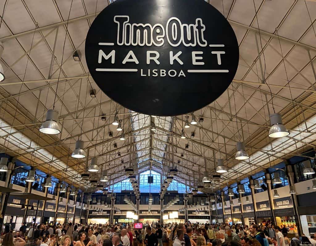 The interior of Time Out Market in Lisbon, Portugal, bustling with people. The market features high ceilings, hanging lights, and numerous food stalls lining the sides. This cafeteria is a high end eatery not to be missed on your 4 days in Lisbon.