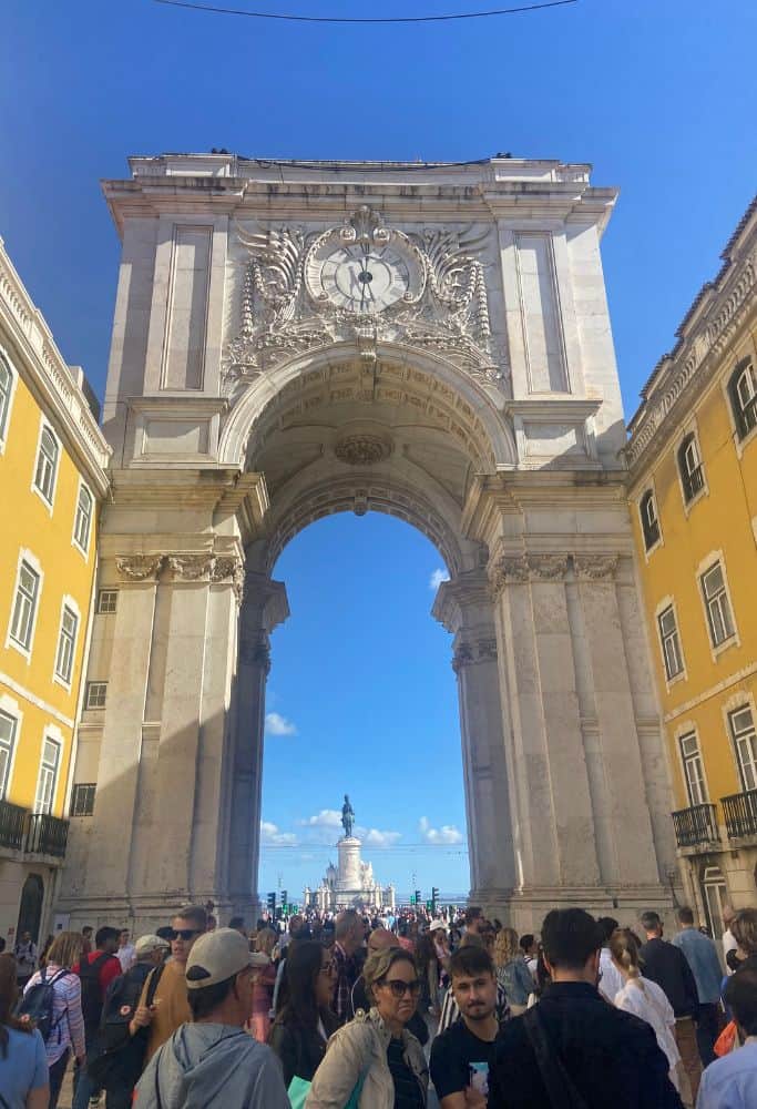 The Arco da Rua Augusta in Lisbon, Portugal, framed by yellow buildings. The archway is adorned with intricate carvings and a clock at the top. Below the arch, a crowd of people is visible, with a statue in the distance.