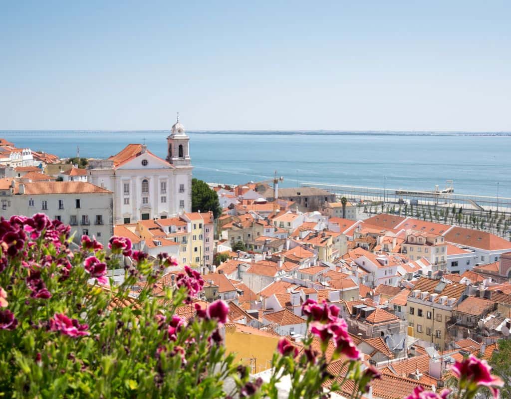 A view of Lisbon, Portugal, from a high vantage point, showing red-tiled rooftops and a white church near the water. The foreground includes vibrant purple flowers, and the sea stretches out in the background.