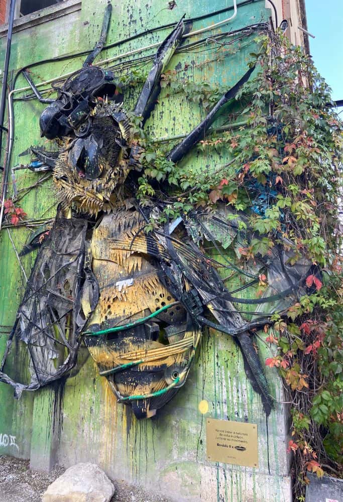 A large sculpture of a bee made from recycled materials, mounted on a green wall with some climbing plants. The sculpture is detailed and incorporates various textures and colors, including black, yellow, and green. The wall is partially covered with ivy and electrical cables. The artwork is located in LX Factory in Lisbon, Portugal.