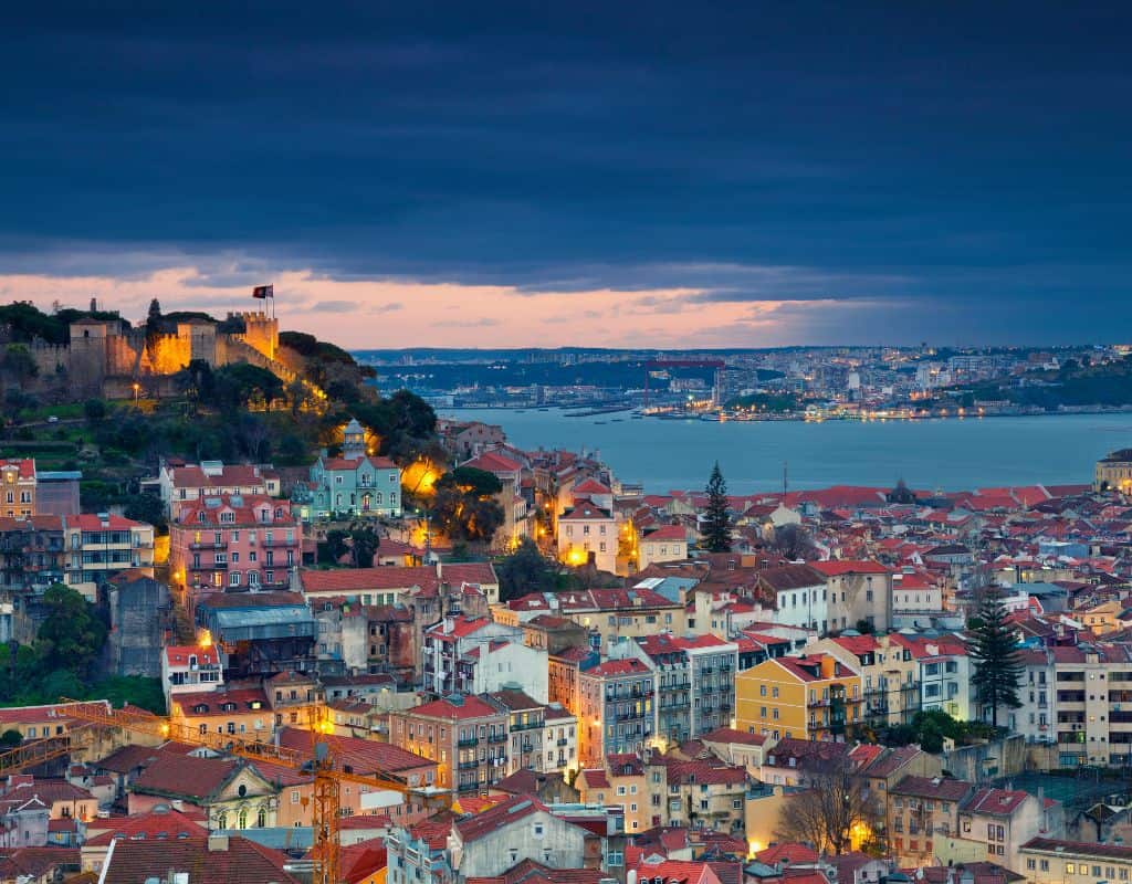 A panoramic view of Lisbon, Portugal, at dusk, featuring the illuminated Castelo de São Jorge on a hill. The cityscape is a mix of red-tiled rooftops and modern buildings, with the Tagus River in the background. This is one of the great views you'll see on a 4 days in Lisbon.