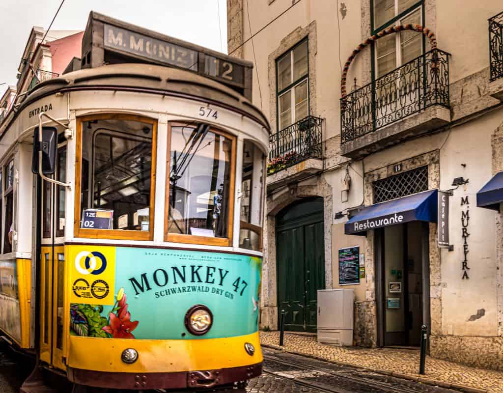 A yellow tram marked with "M. Moniz" navigating a narrow street in Lisbon, Portugal. The tram passes by buildings with wrought iron balconies and a restaurant, highlighting the charm to be seen on a 4 days in Lisbon.