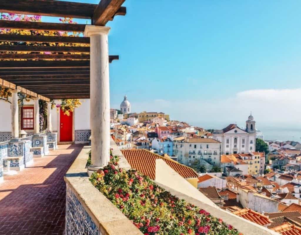 A panoramic view from a view point in Alfama, in Lisbon, Portugal, featuring a pergola with columns and tiled benches. The scene overlooks a picturesque cityscape with rooftops, churches, and the distant sea.