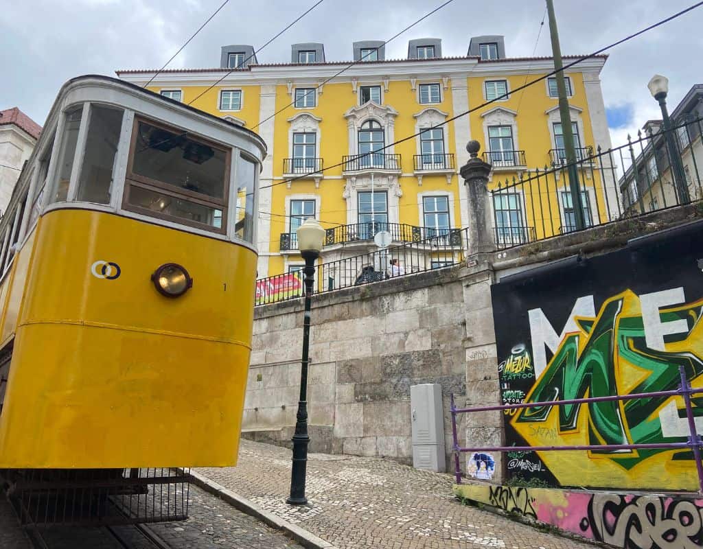 A close-up of the elevador da Gloria in Lisbon, Portugal, against the backdrop of a bright yellow building with ornate architectural details. Graffiti decorates the wall next to the tram.