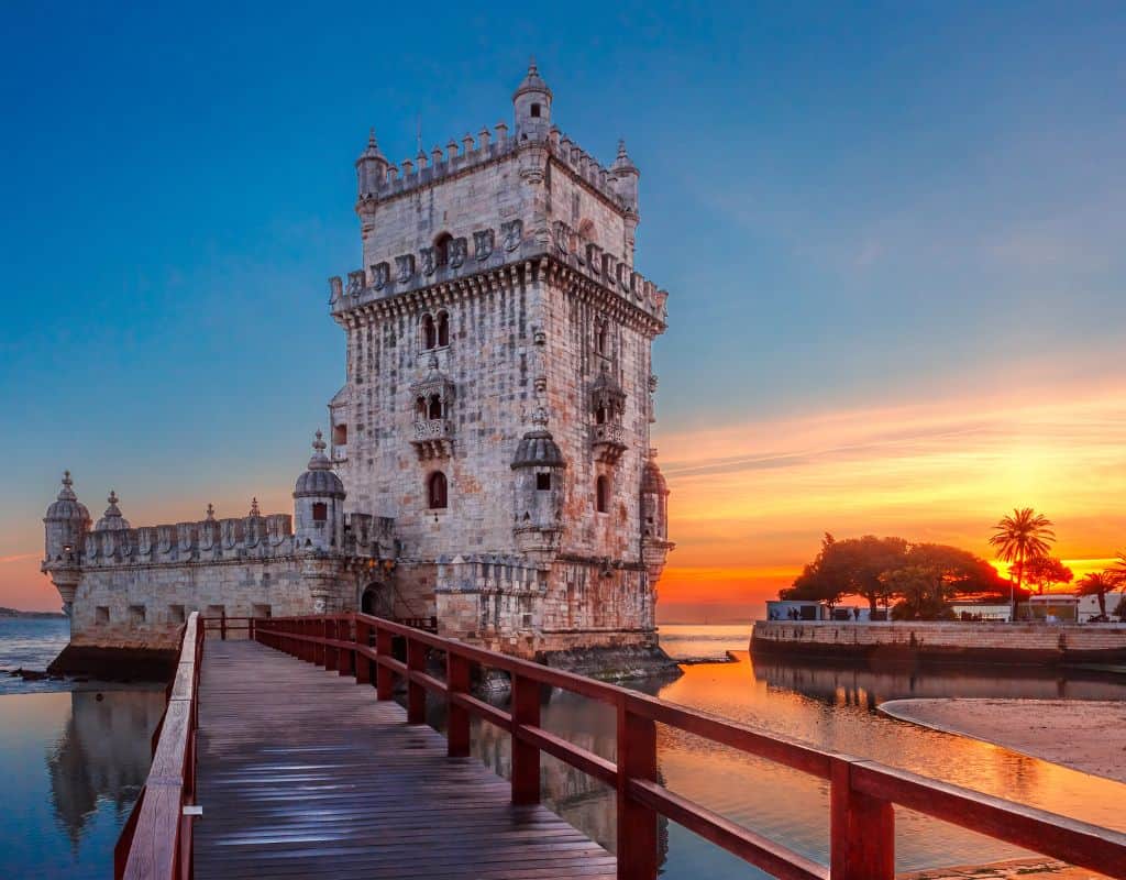 Belém Tower in Lisbon, Portugal, at sunset. The historic tower stands on the water's edge, connected to the shore by a wooden walkway. The sky is painted with warm hues of orange and pink.