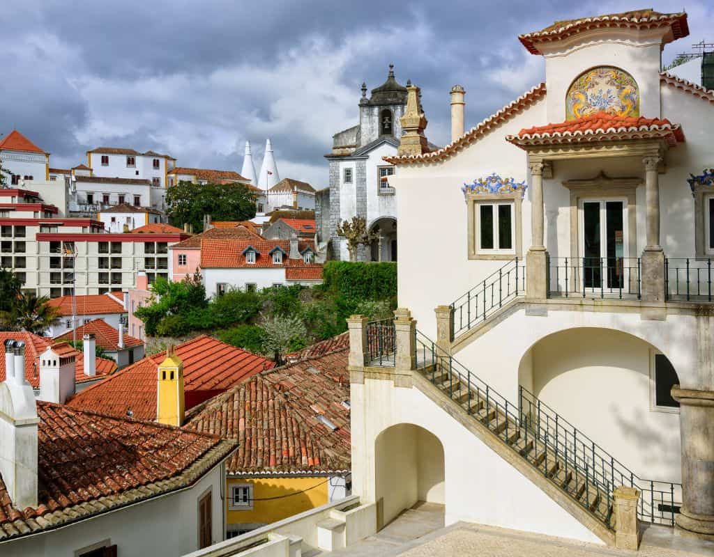 A scenic view of Sintra, Portugal, showcasing traditional architecture with red-tiled rooftops and ornate facades. The background features a church and  twin white chimneys from the Nacional Royal Palace, under a cloudy sky. This is a great day trip from Lisbon which can easily be planned into your 4 days in Lisbon.