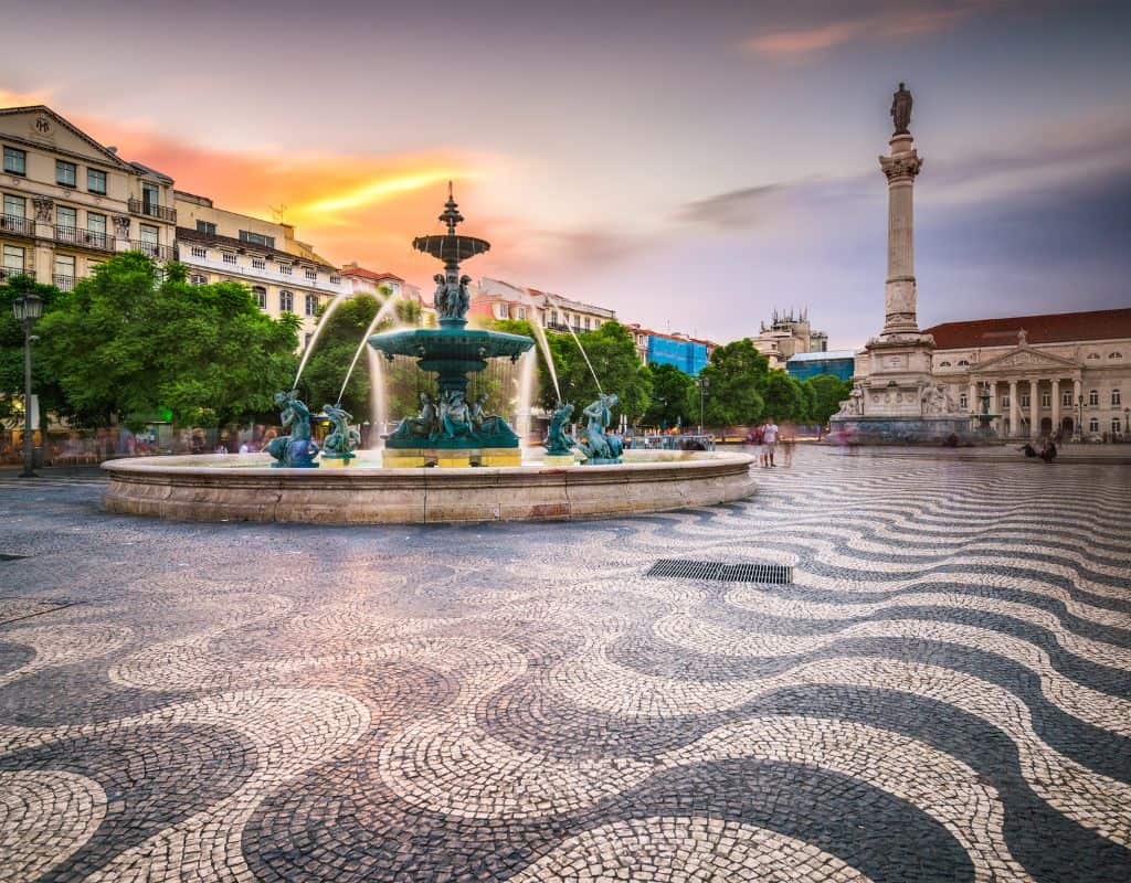 Rossio Square in Lisbon, Portugal, at sunset. The square features a large fountain surrounded by statues, with water spraying into the air. The ground is paved with intricate black and white patterns, and the background includes historic buildings and a tall monument.