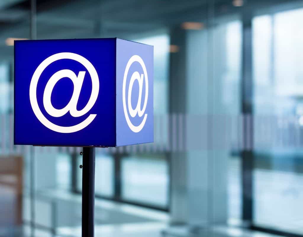 A glowing "@" symbol on a blue cube sign in an airport setting, representing digital communication or internet access.