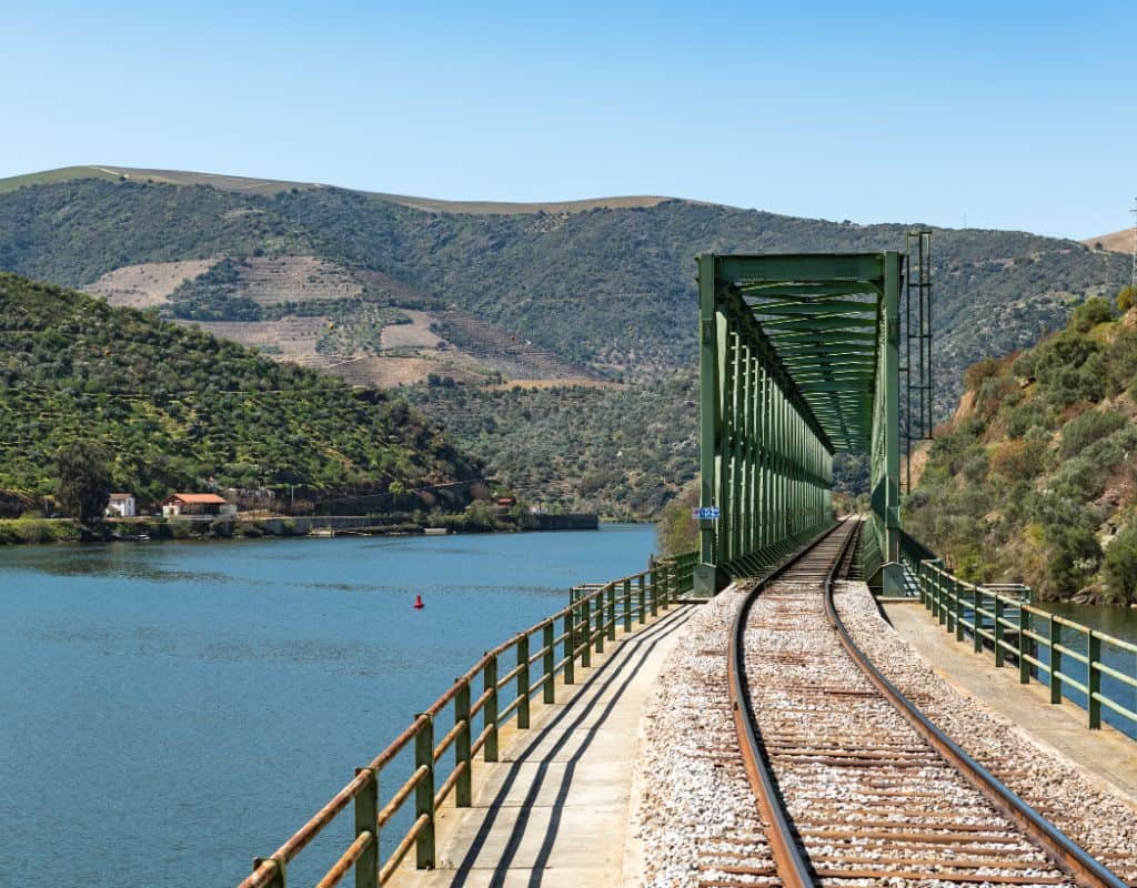 A steel bridge carrying a train track over a blue river, surrounded by lush green hills on a sunny day. This spot is part of the picturesque Porto to Douro Valley by Train experience.