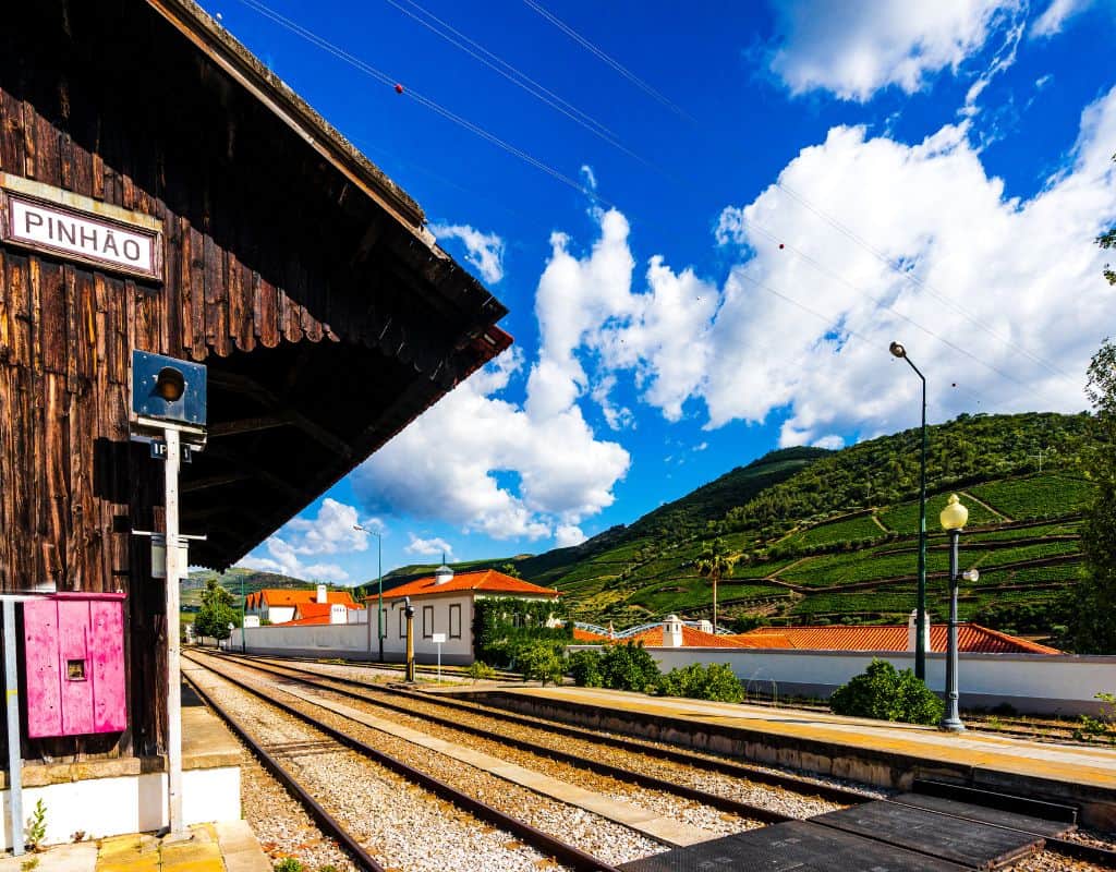 The rural train station named "Pinhão" with railway tracks stretching into the distance, surrounded by hills and vineyards under a bright blue sky. This is the place to stop on your trip from Porto to Douro Valley by train.