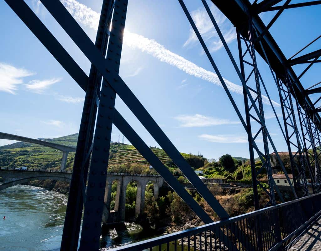 A steel bridge carrying a train track over a blue river, surrounded by lush green hills. This spot is part of the picturesque Porto to Douro Valley by Train experience.