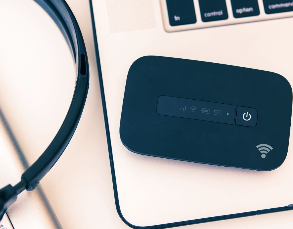 A portable Wi-Fi hotspot device lies on top of a laptop beside some headphones, possibly used with a Portugal SIM Card for on-the-go connectivity.