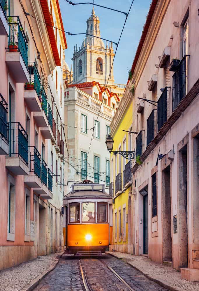 A narrow street in Portugal with traditional buildings, featuring a classic yellow tram moving along the tracks towards the camera. The tram is framed by balconies and an ornate church spire in the background.