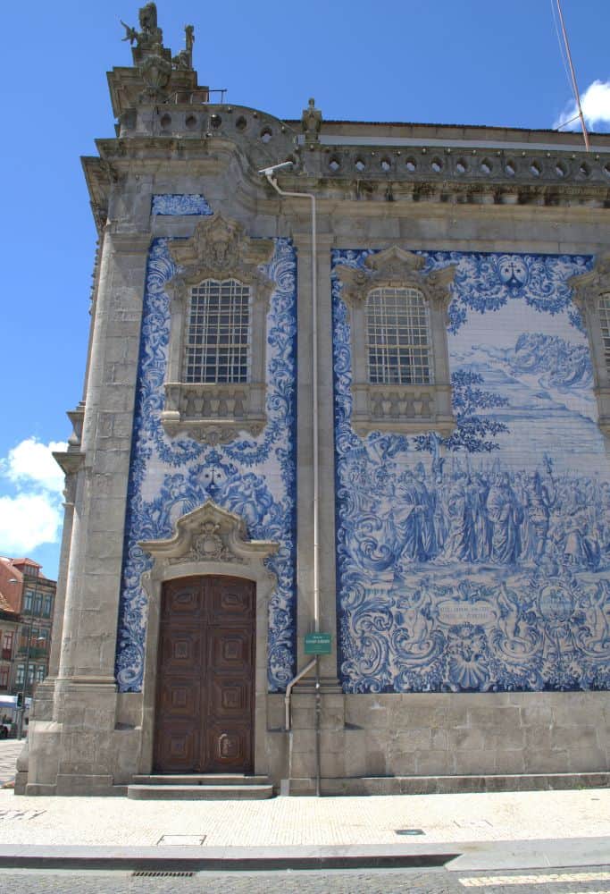 A close-up of a historic Portuguese church adorned with intricate blue-and-white tiles. The grand entrance features wooden doors and sculpted stone details.
