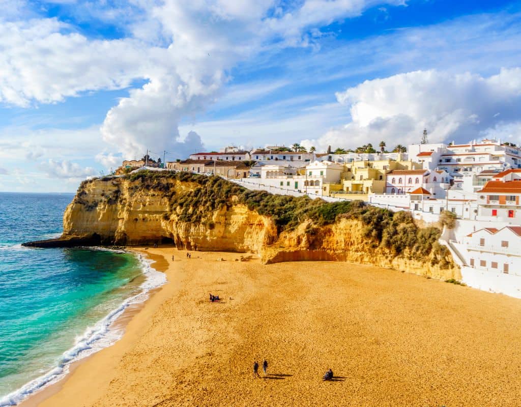 The sunlit beach and village of Carvoeiro framed by golden cliffs, with a small village perched on the edge of the cliff, overlooking the sandy shore and ocean waves.