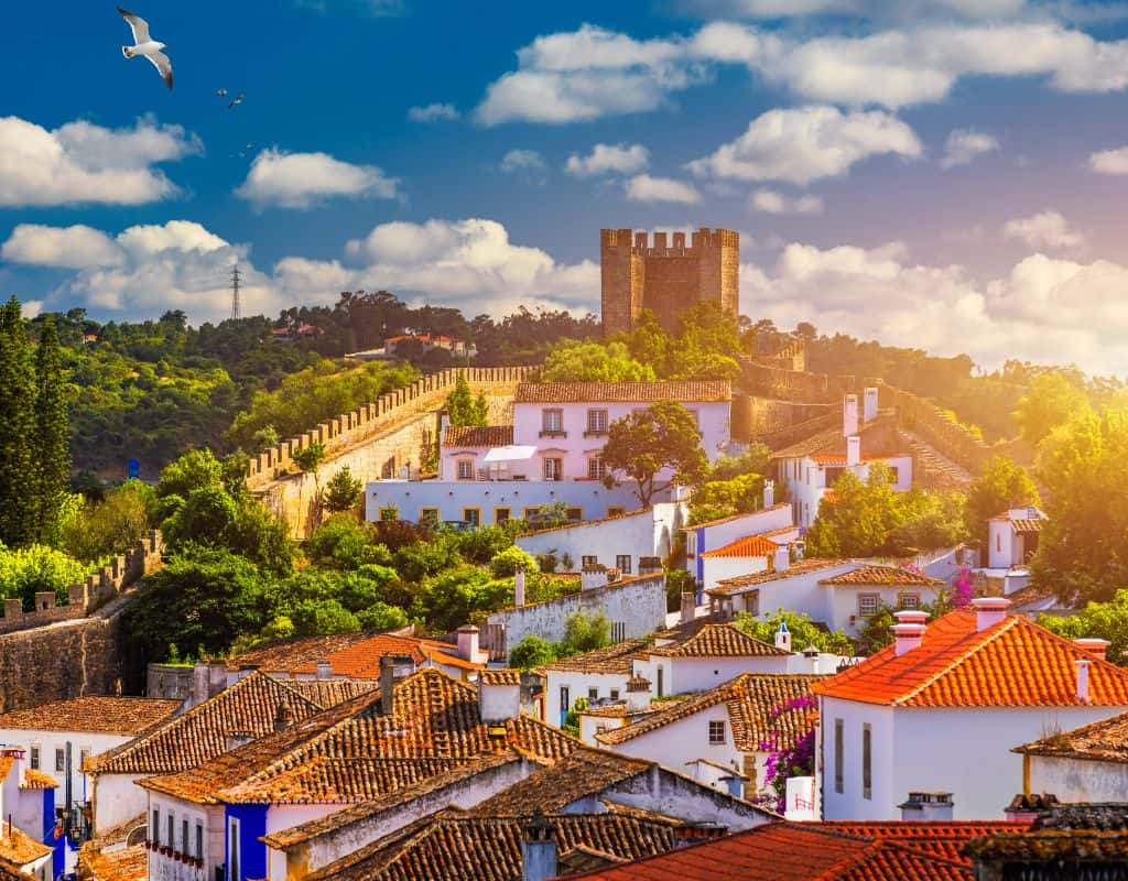 The charming medieval town of Obidos in Portugal with whitewashed houses and terracotta rooftops, surrounded by a historic stone wall. A castle overlooks the town from a hilltop in the distance.